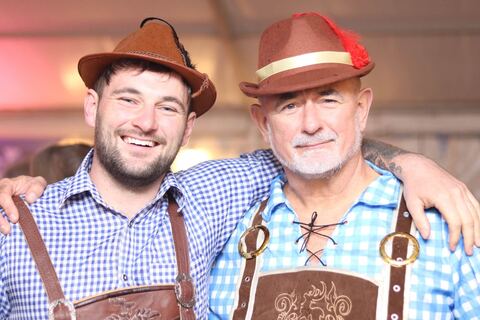 Two men in feathered Alpine hats and lederhosen braces, arms round each other, grinning.