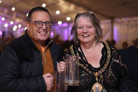 Two guests smile with their steins under festoon lights, one wearing a chain of office.